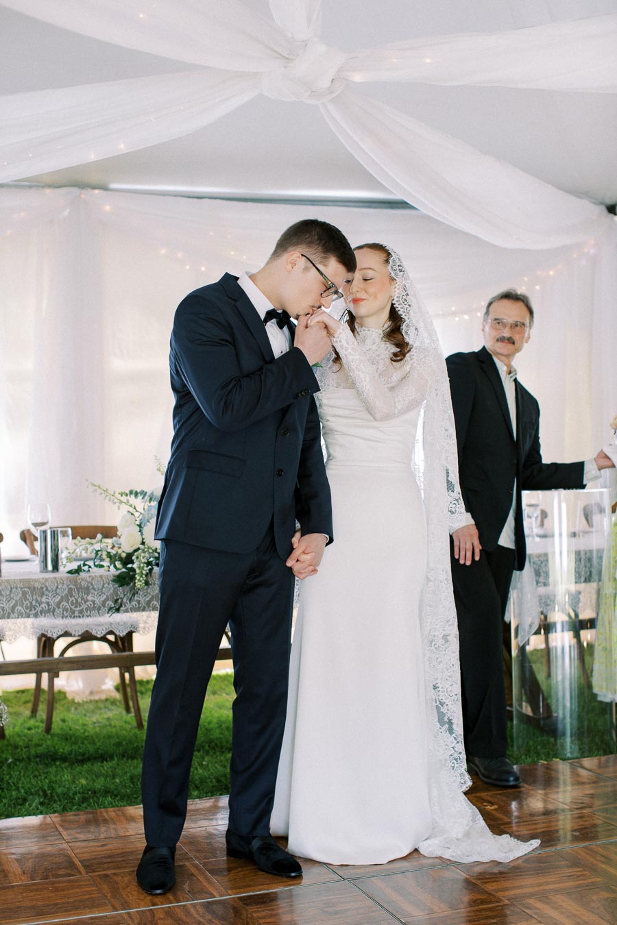 A bride and groom share a tender moment during their wedding ceremony, with the groom gently kissing the bride's hand. Both are dressed in elegant formal attire under a beautifully draped tent, accompanied by a warmly smiling officiant in the background.