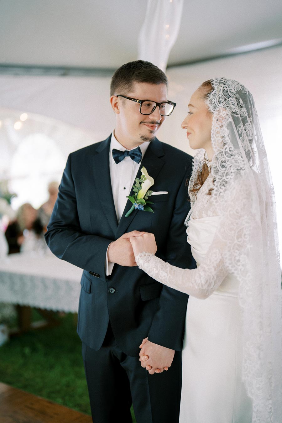 A bride and groom smiling at each other, holding hands during their wedding ceremony under a decorated tent, with the bride in a lace veil and the groom in a navy suit.