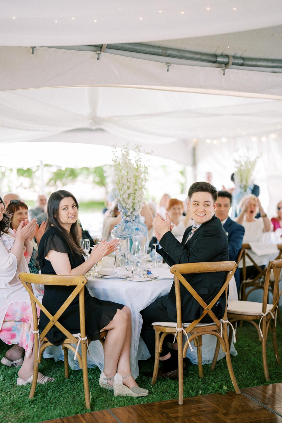 A group of elegantly dressed wedding guests clapping and smiling while seated at a beautifully decorated table under a white tent, featuring wooden chairs and floral centerpieces.