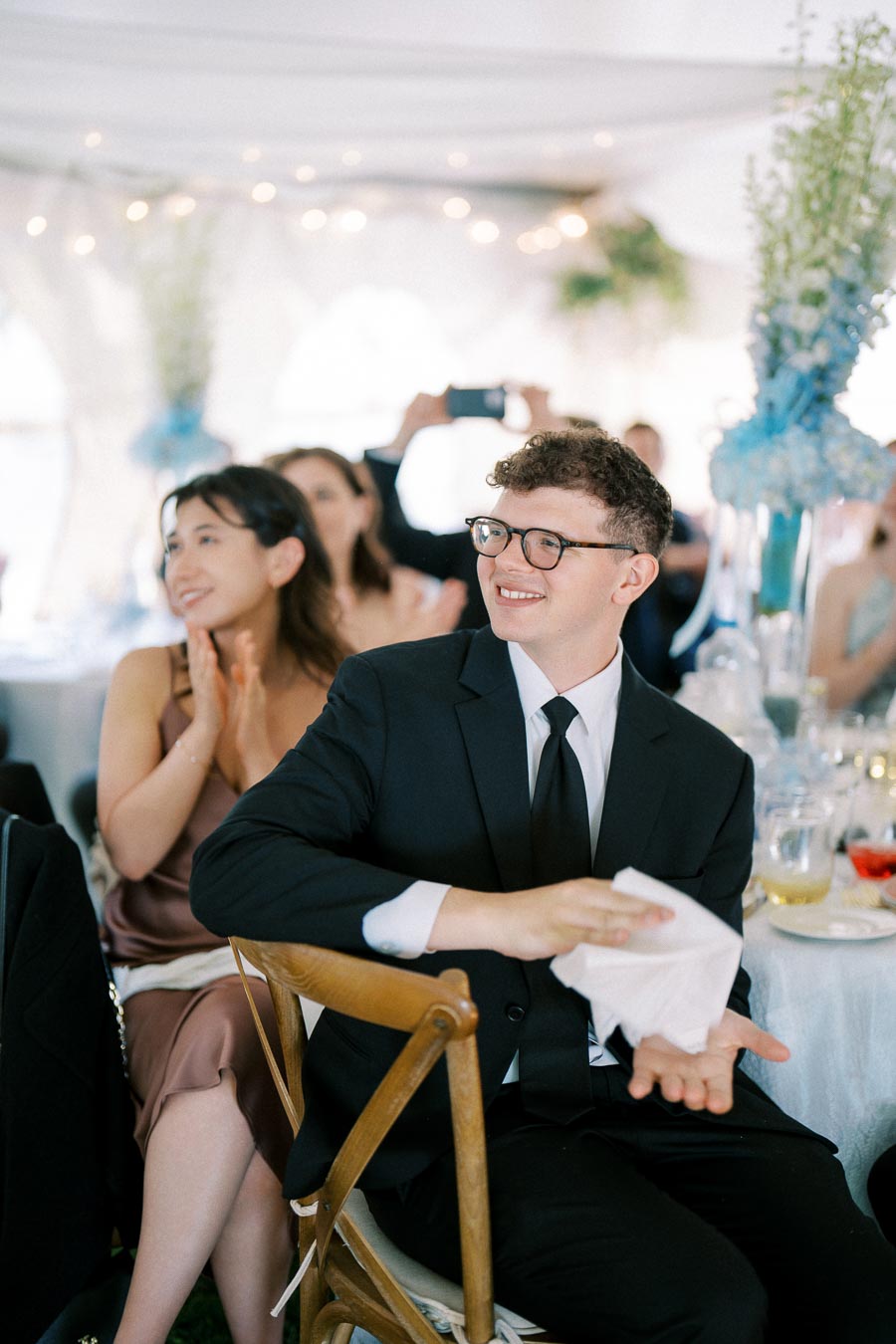A young man in a black suit and glasses sits smiling and clapping at a formal event, surrounded by applauding guests in an elegantly decorated venue.