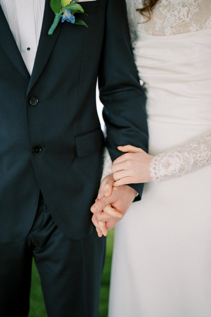 A bride and groom holding hands, dressed in a white lace wedding dress and a dark suit with a floral boutonniere.