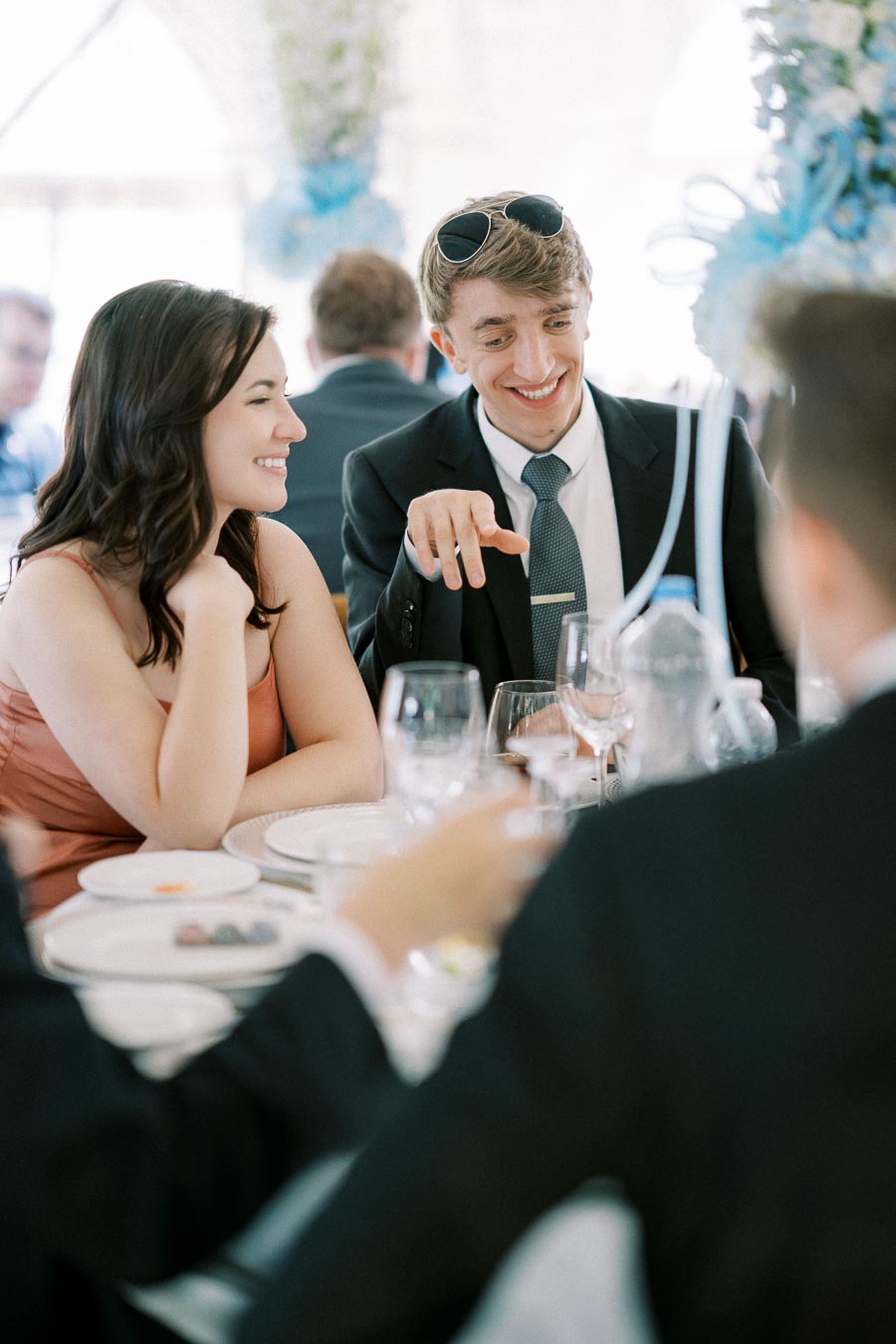 Two people smiling and enjoying a conversation at a formal event, elegantly dressed and seated at a table with plates and wine glasses.