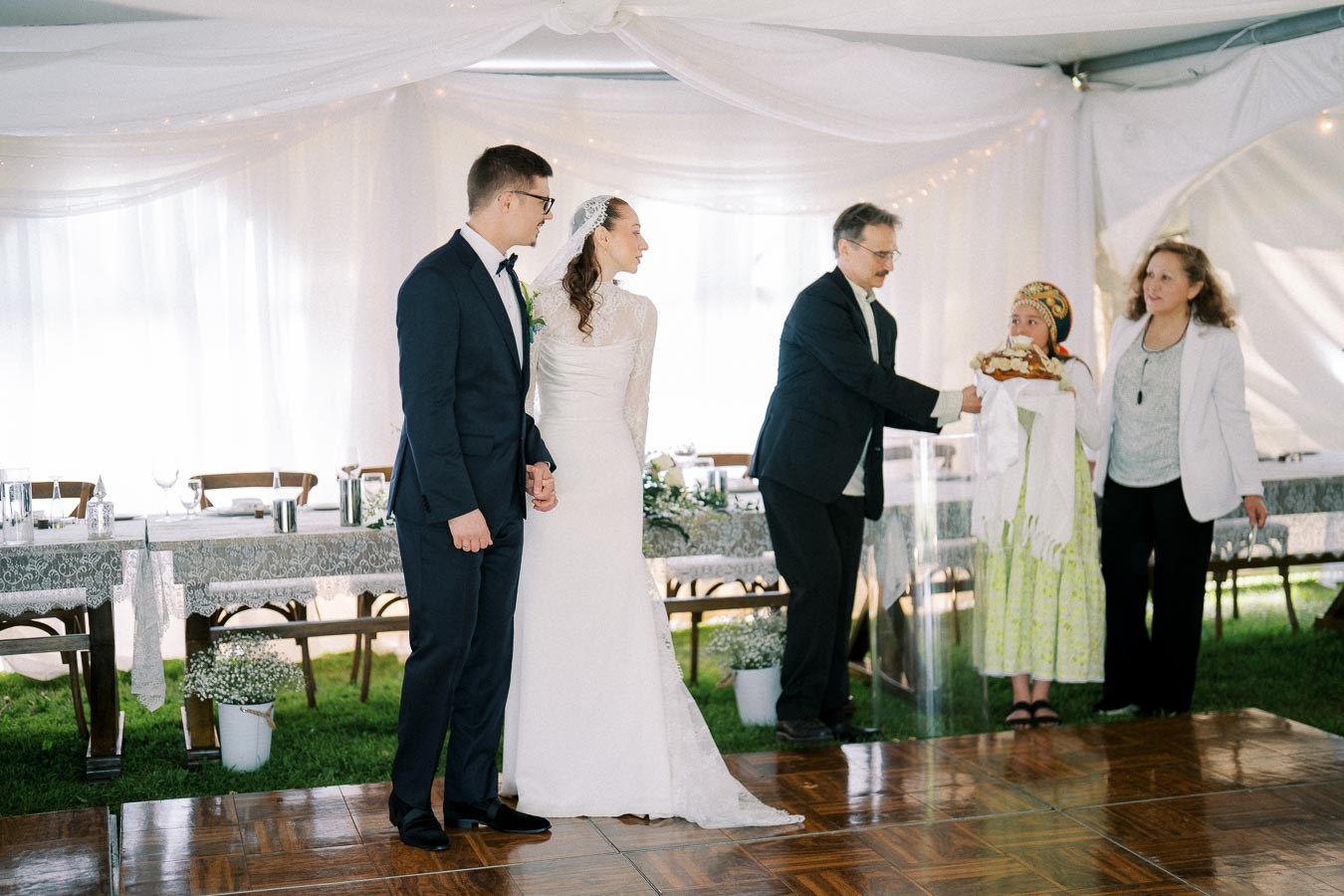 A bride and groom stand together during a traditional wedding ceremony with family members under a white canopy, featuring a beautifully decorated table and guests in formal attire.
