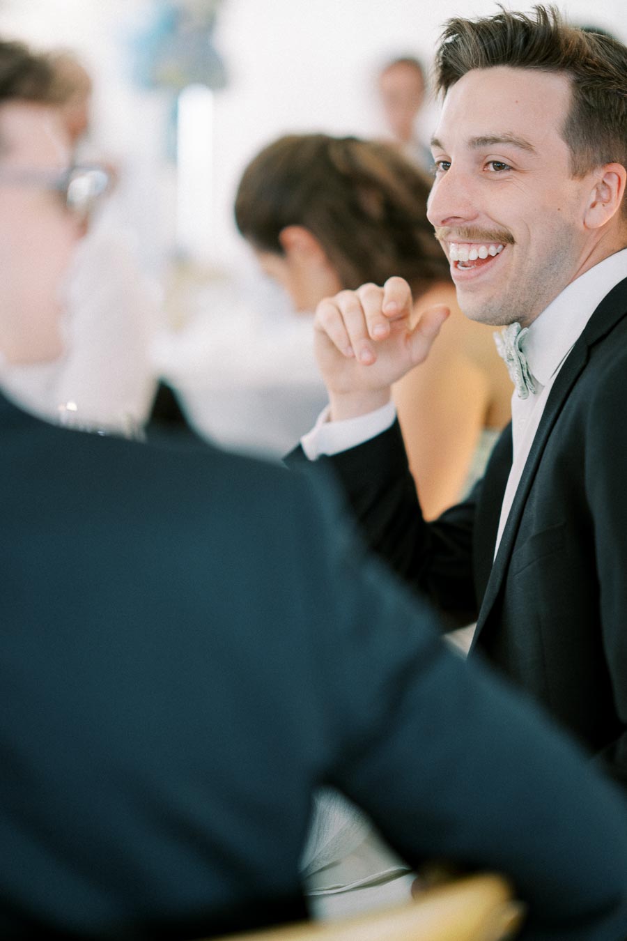 A man in a suit smiling and conversing at a formal event, with blurred guests in the background.