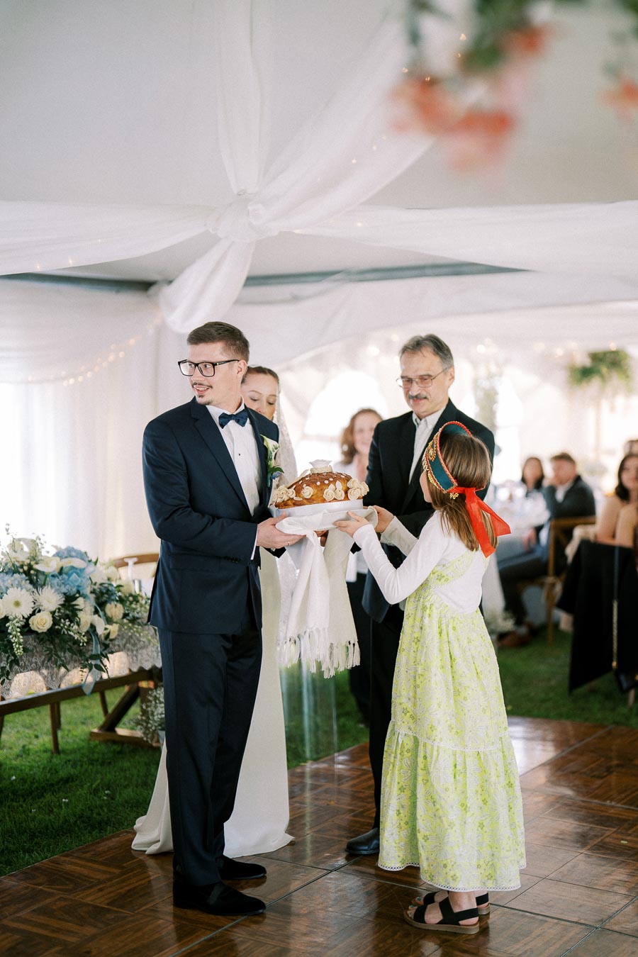 Wedding ceremony with groom in suit receiving traditional bread from young girl in festive dress, symbolizing welcoming and cultural customs.