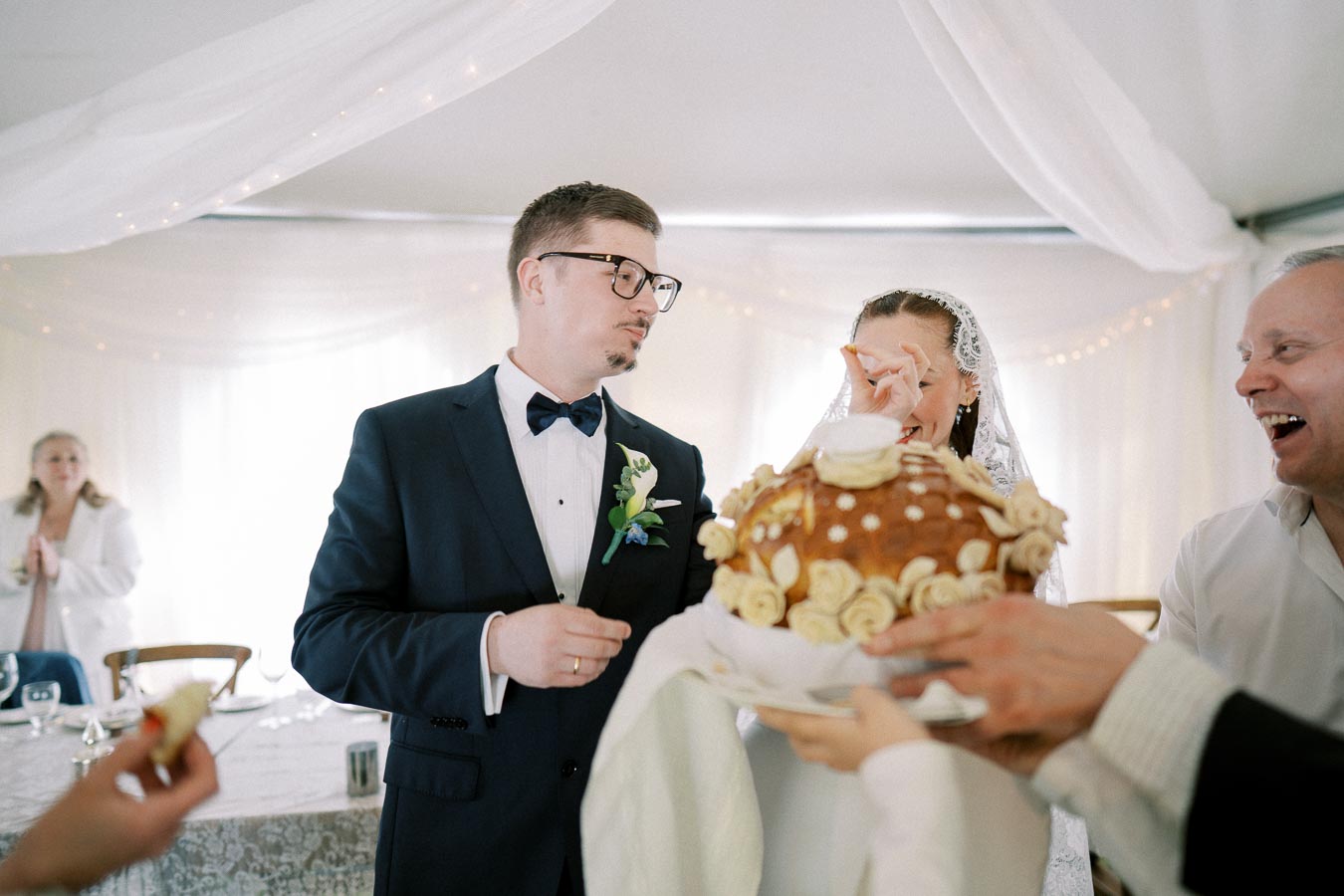 A bride and groom participate in a traditional ceremony, breaking bread adorned with floral decorations, surrounded by wedding guests in a decorated venue.