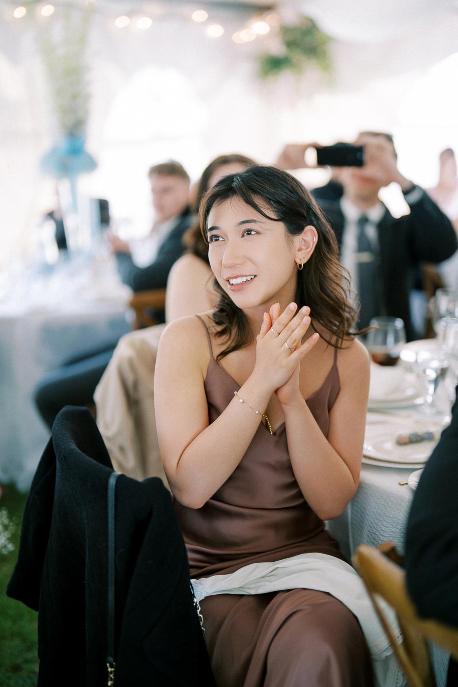 A young woman in a brown dress smiling and applauding at a formal event, seated at a table set with elegant dinnerware, surrounded by people in a softly lit environment.