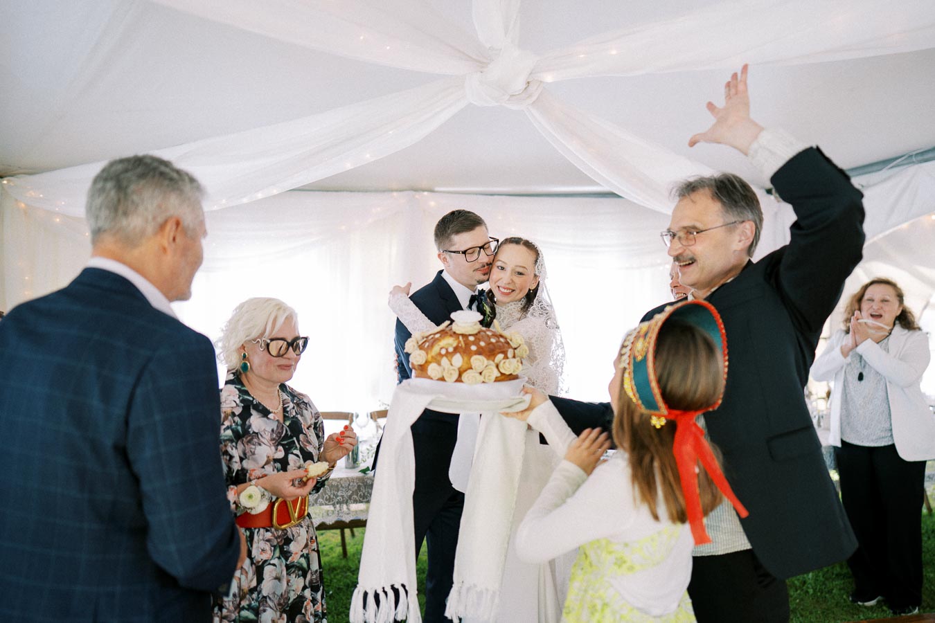 A joyful wedding celebration with a bride and groom smiling surrounded by guests in a decorated tent, holding a traditional decorative bread.