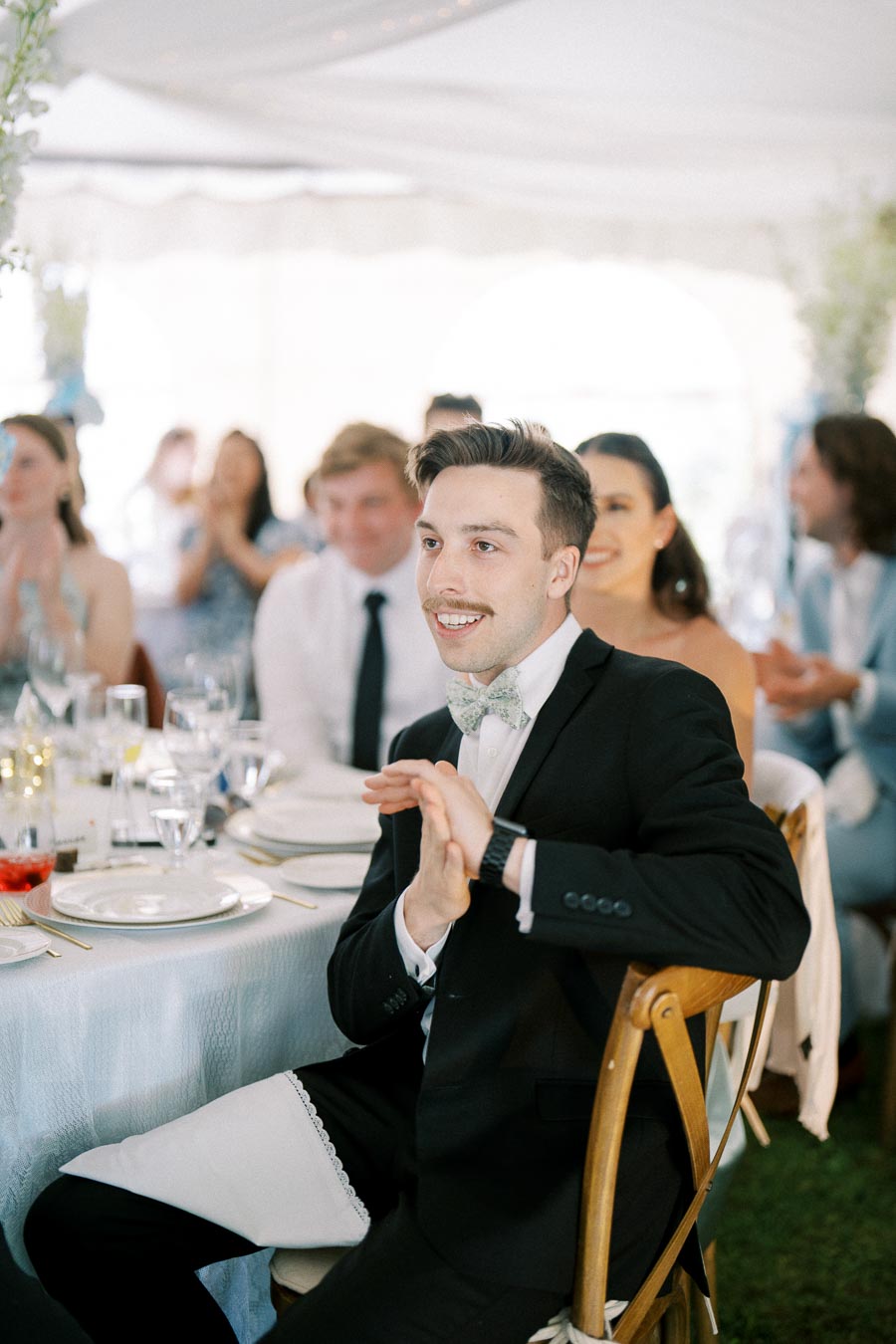 A well-dressed man in a black suit and bow tie smiles while sitting at an elegantly set table during a formal event. Other guests are visible in the background, clapping and enjoying the occasion inside a beautifully decorated tent.