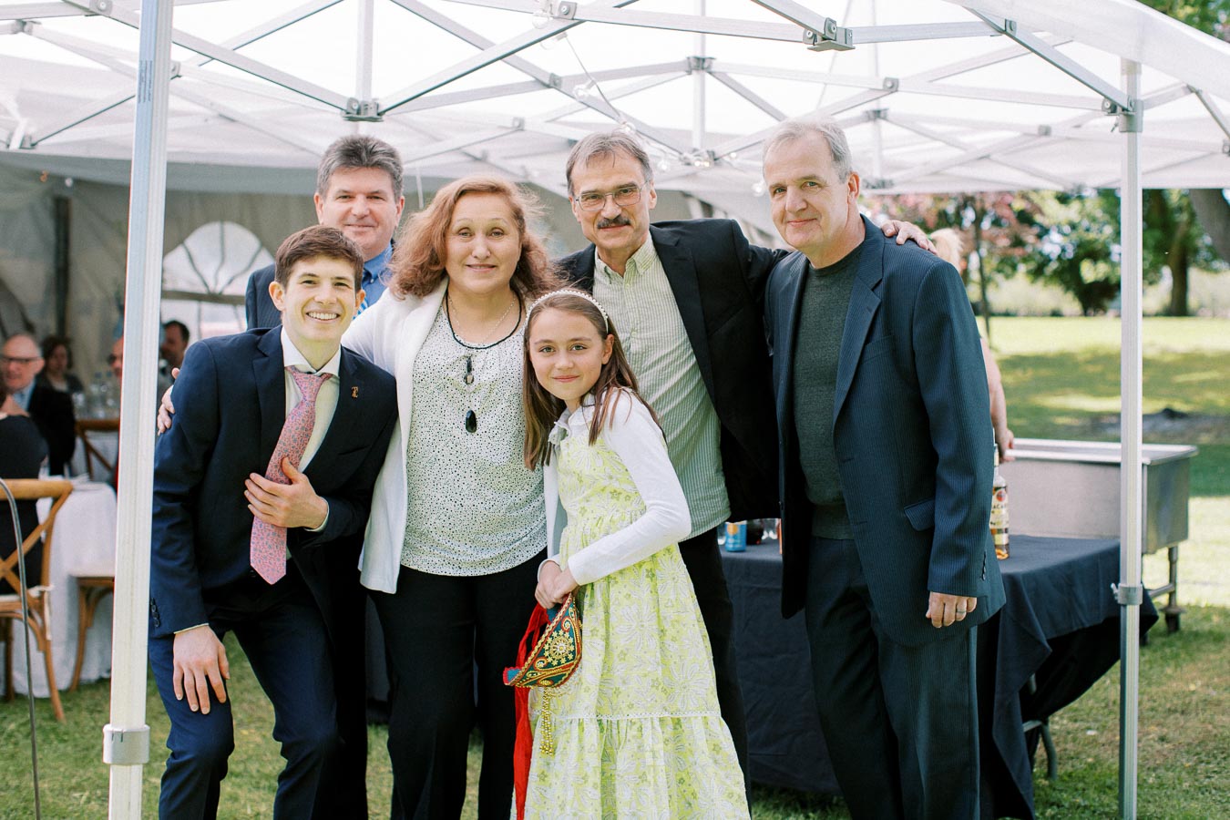 A group of people smiling and posing together at an outdoor event under a white canopy, including a young girl in a floral dress and adults in formal attire, with a green lawn and trees in the background.