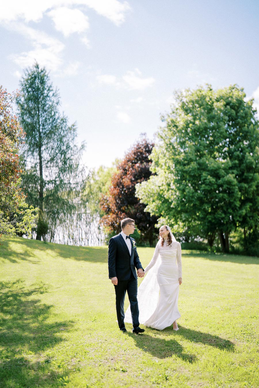 A bride and groom hold hands walking through a lush green park on a sunny day, surrounded by vibrant trees, capturing a beautiful wedding moment outdoors.