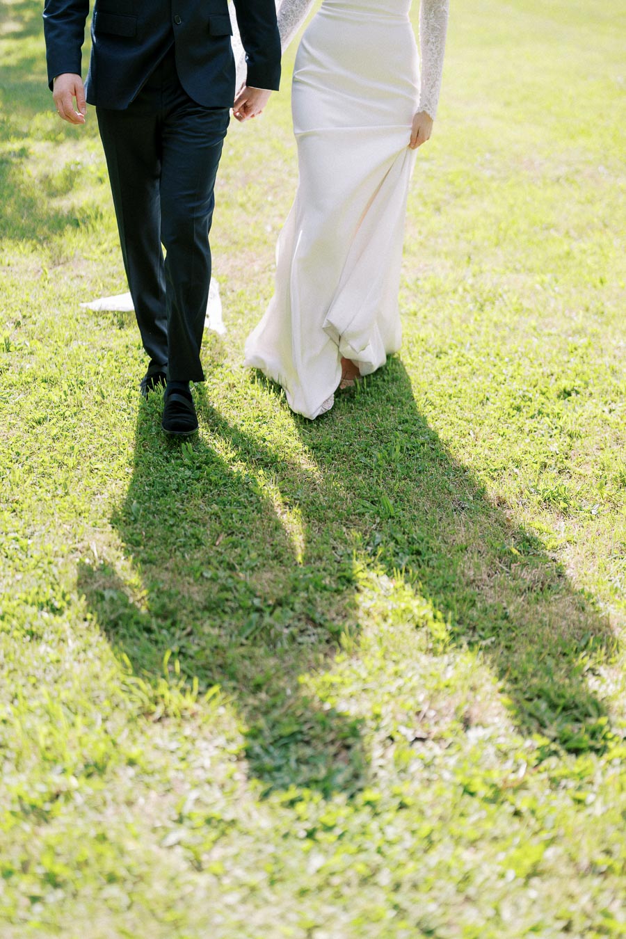 A bride in a white wedding dress and groom in a black suit walking on green grass, casting long shadows in sunlight.