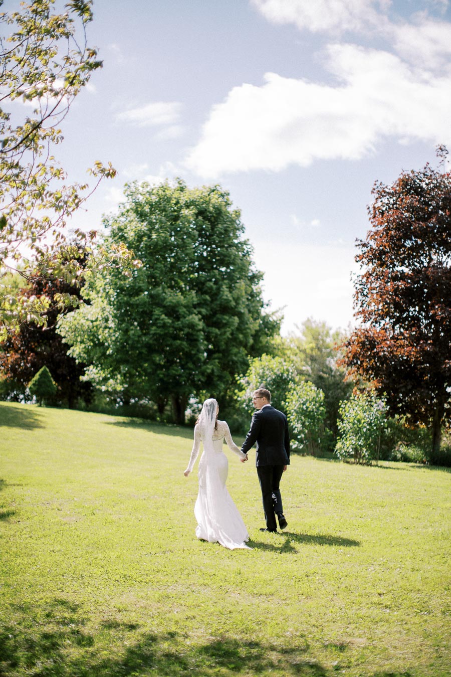A bride and groom holding hands walking on a grassy field surrounded by lush, green trees under a clear blue sky.