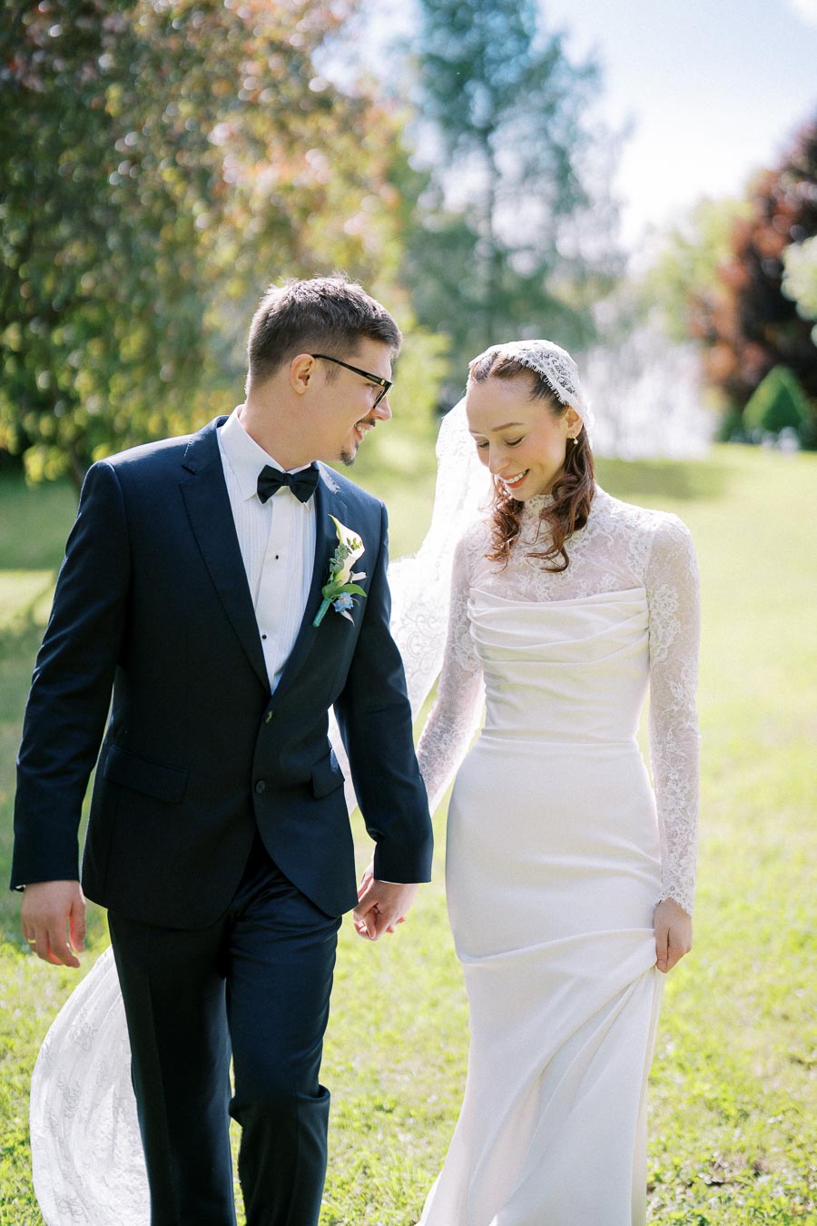A happy couple in elegant wedding attire walks hand-in-hand on a sunny day in a lush green garden, with the bride wearing a lace dress and veil and the groom in a black suit and bow tie.