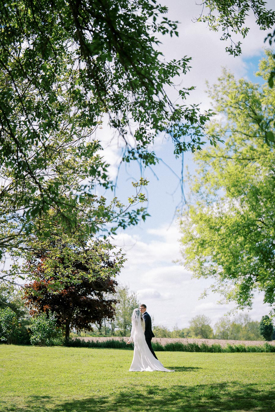 A bride and groom standing in a lush green field under a canopy of trees, with a picturesque sky in the background, captures a beautiful outdoor wedding moment.