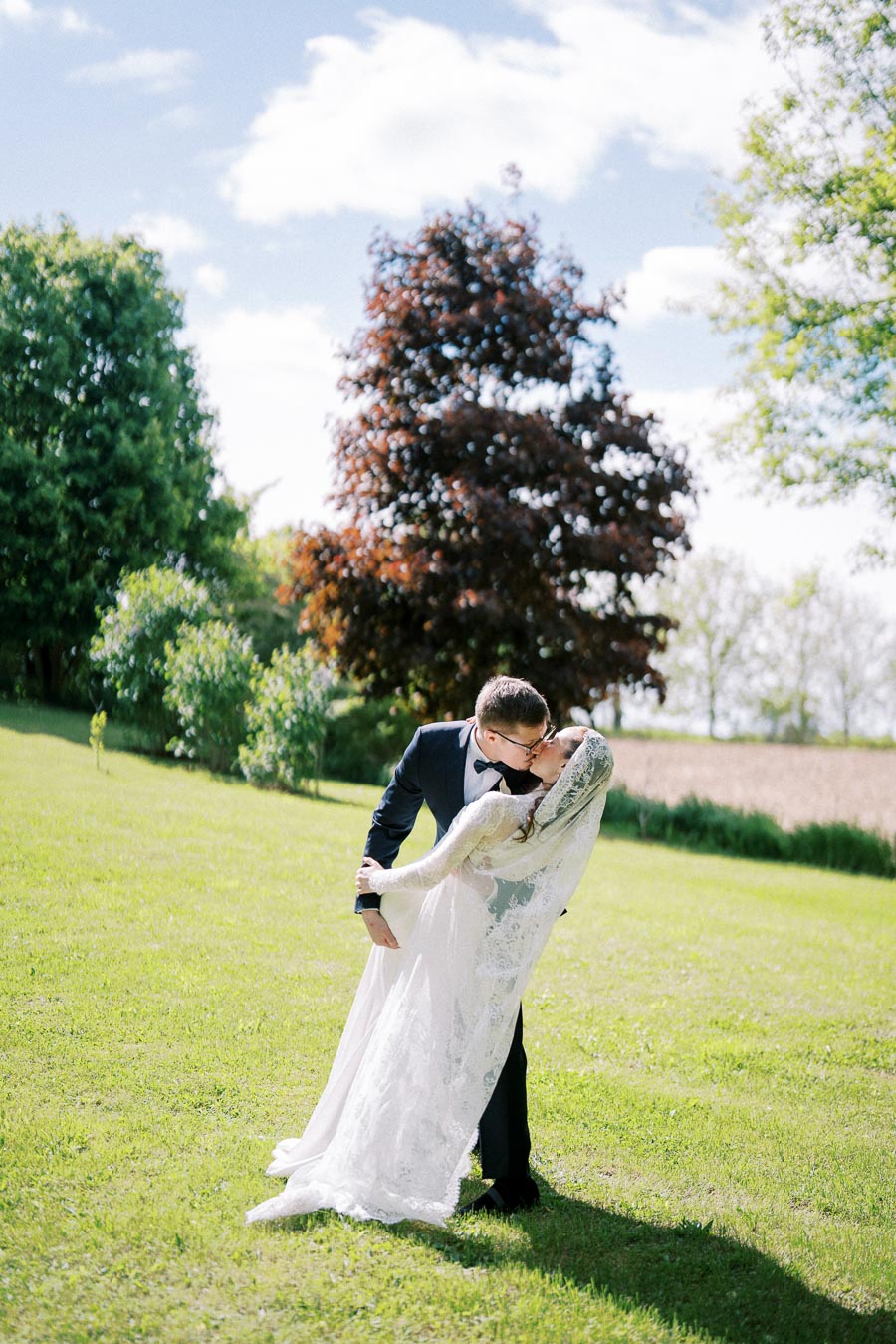 A bride and groom share a romantic kiss on a green lawn, surrounded by lush trees, under a sunny sky. The bride is wearing a lace dress and veil, and the groom is in a blue suit with a bow tie. Perfect for a picturesque outdoor wedding setting.