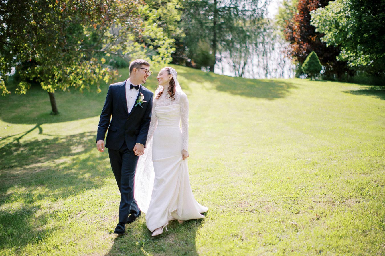 Happy bride and groom walking hand in hand on a sunny day in a lush green park, wearing elegant wedding attire with a backdrop of trees and natural scenery.