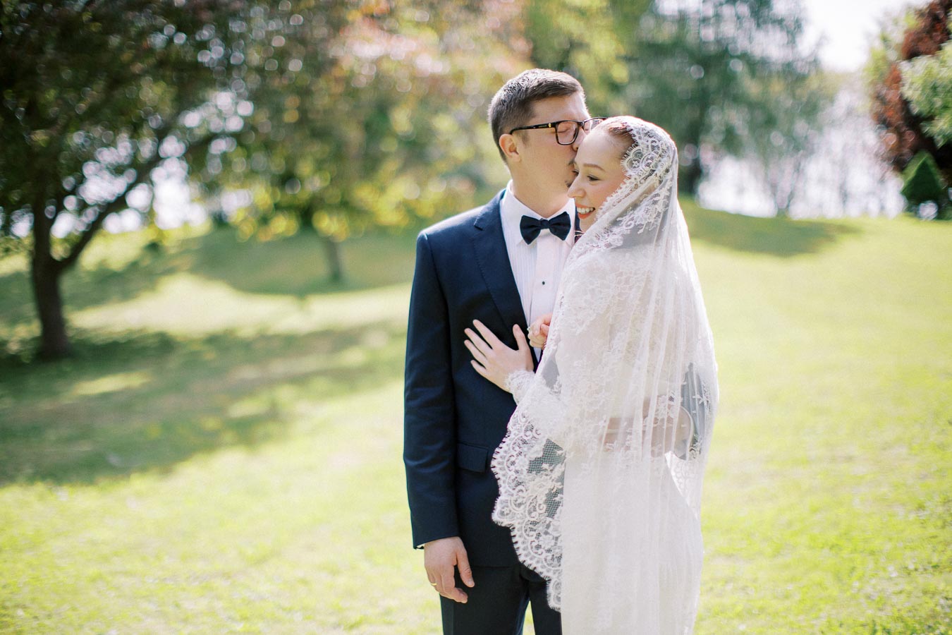 Outdoor wedding scene with a groom in a navy blue suit and bow tie kissing his bride, who is wearing a lace veil, in a sunlit garden with trees in the background.