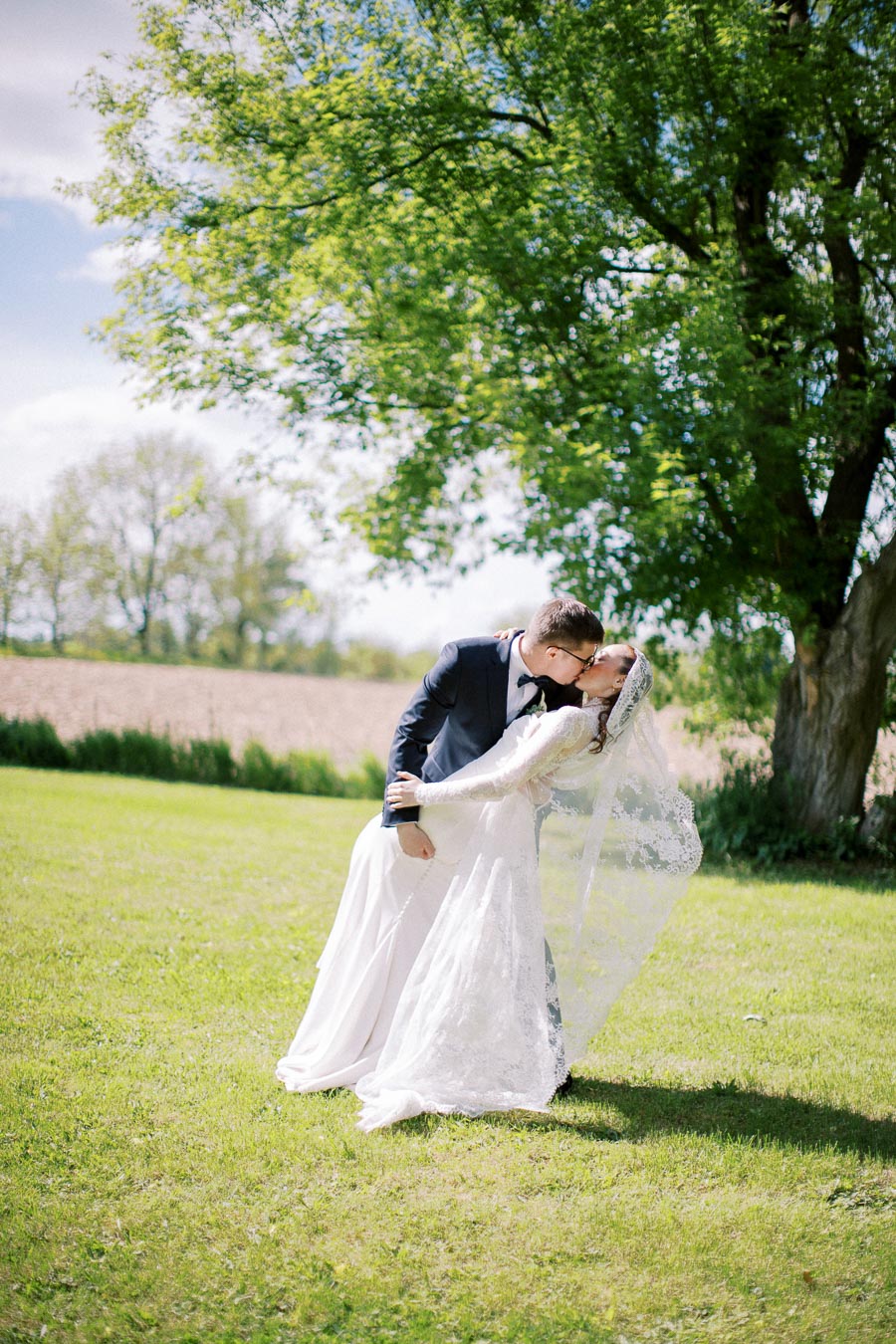 A bride in a white lace gown and groom in a navy suit share a romantic kiss under a large tree on a sunny day in a lush green field.