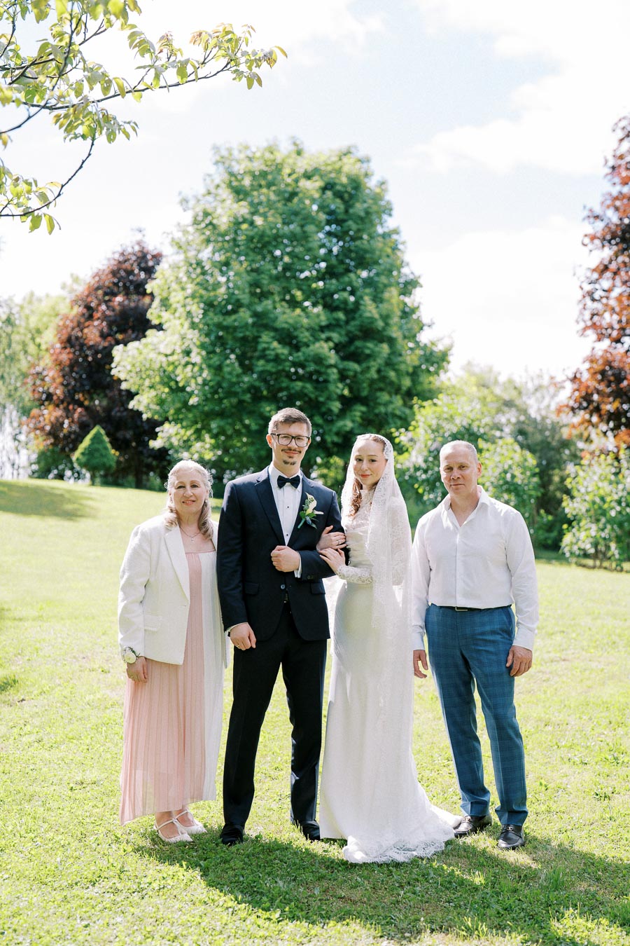 Wedding couple posing with their parents in a scenic garden setting. The groom is wearing a black tuxedo, and the bride is in a white lace gown. The parents are dressed in formal attire, standing on vibrant green grass with lush trees in the background.
