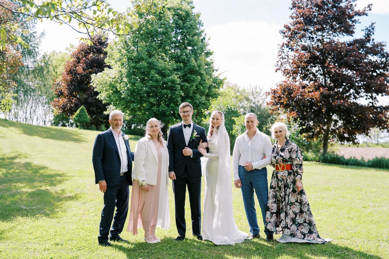 A wedding photo of a happy couple surrounded by family in an outdoor setting with lush green trees and a sunny sky. The bride is wearing a white gown, and the groom is in a black suit. The family is dressed in formal attire, smiling in celebration.