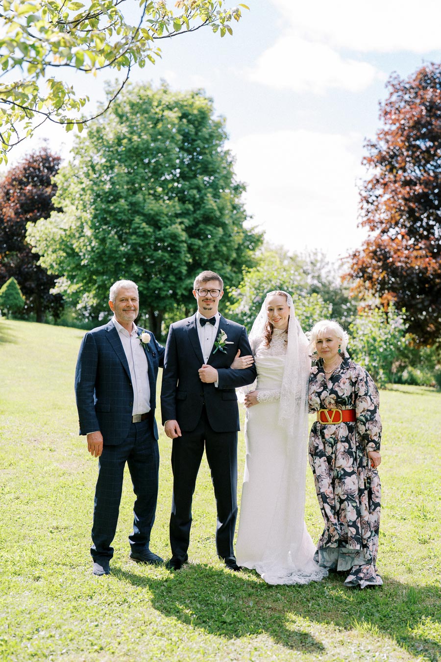 A wedding photo featuring a groom in a tuxedo and a bride in a white dress and veil, posing with two older adults in a sunny outdoor park setting with lush green trees in the background.