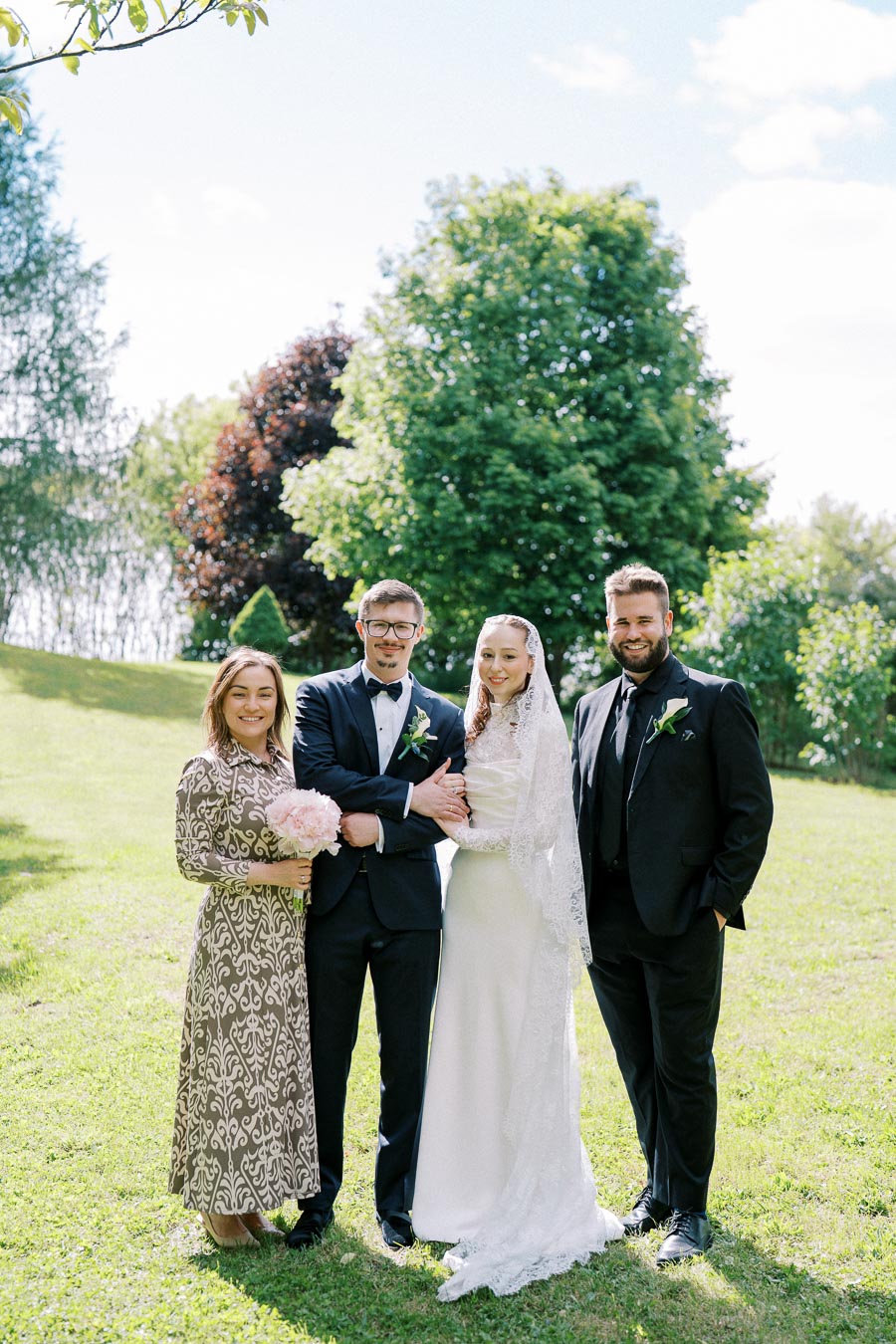 A joyful wedding photo featuring a smiling bride in a white gown and lace veil, standing alongside a groom in a black tuxedo. They are surrounded by two elegantly dressed friends, one holding a bouquet of pink flowers, in a lush green garden on a sunny day.
