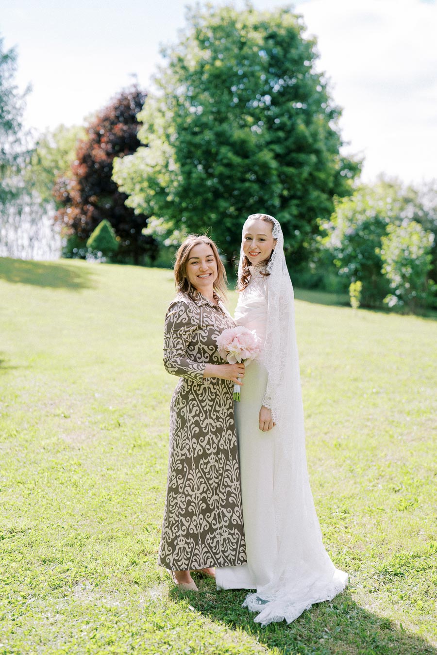 Two women standing on a sunlit grassy lawn, one in a patterned dress and the other in a white bridal gown with lace veil, both holding a bouquet of pink flowers, surrounded by lush green trees.