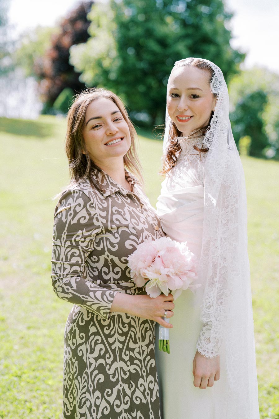 Two smiling women posing outdoors in a lush green park, one wearing a white lace wedding dress and veil, holding a bouquet of pink flowers, representing a joyful wedding celebration.
