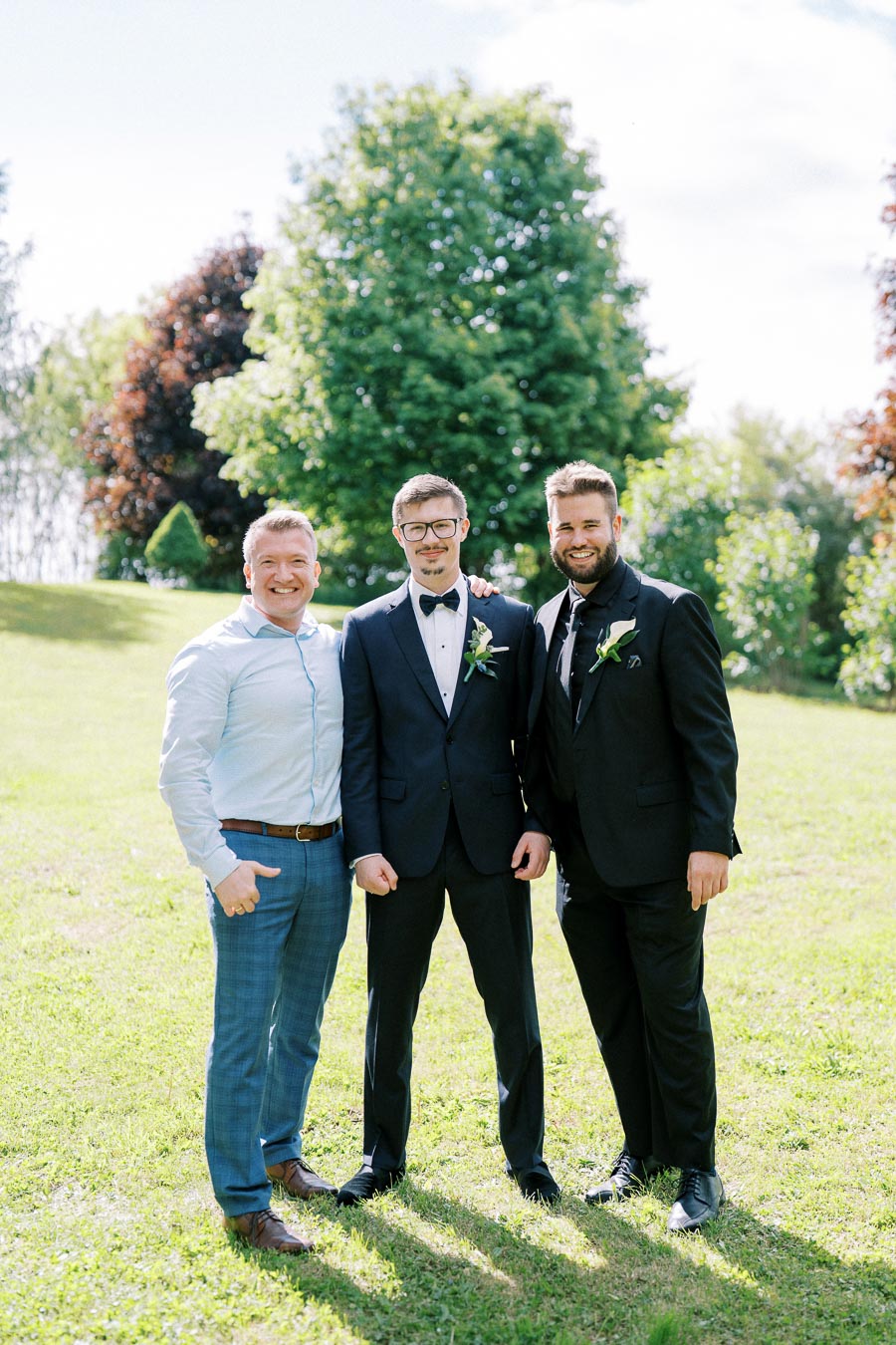 Three men smiling and posing in a sunny park, with one wearing a blue suit and two in black suits with matching boutonnieres, standing on green grass with lush trees in the background.