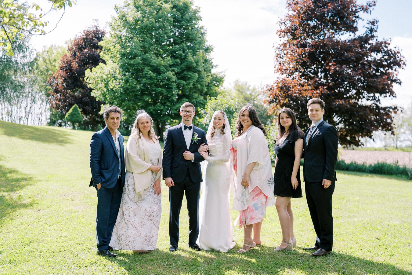 A wedding party posing outdoors with lush green trees in the background, featuring a bride in a white gown, a groom in a black suit, and elegantly dressed family members on a sunny day.