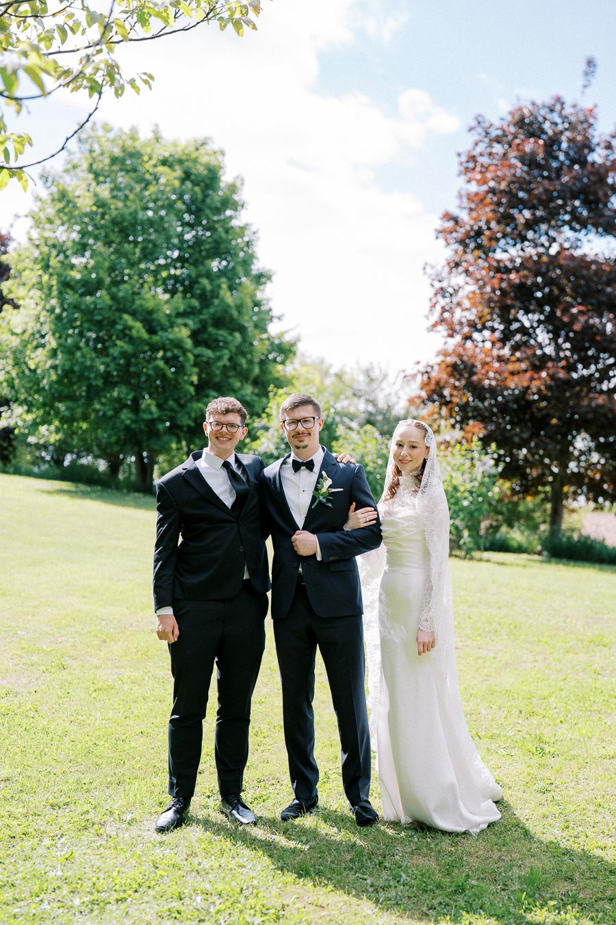 A bride and groom stand with a groomsman on a sunny day in a lush green park, wearing formal wedding attire; trees and a bright blue sky provide a picturesque background.