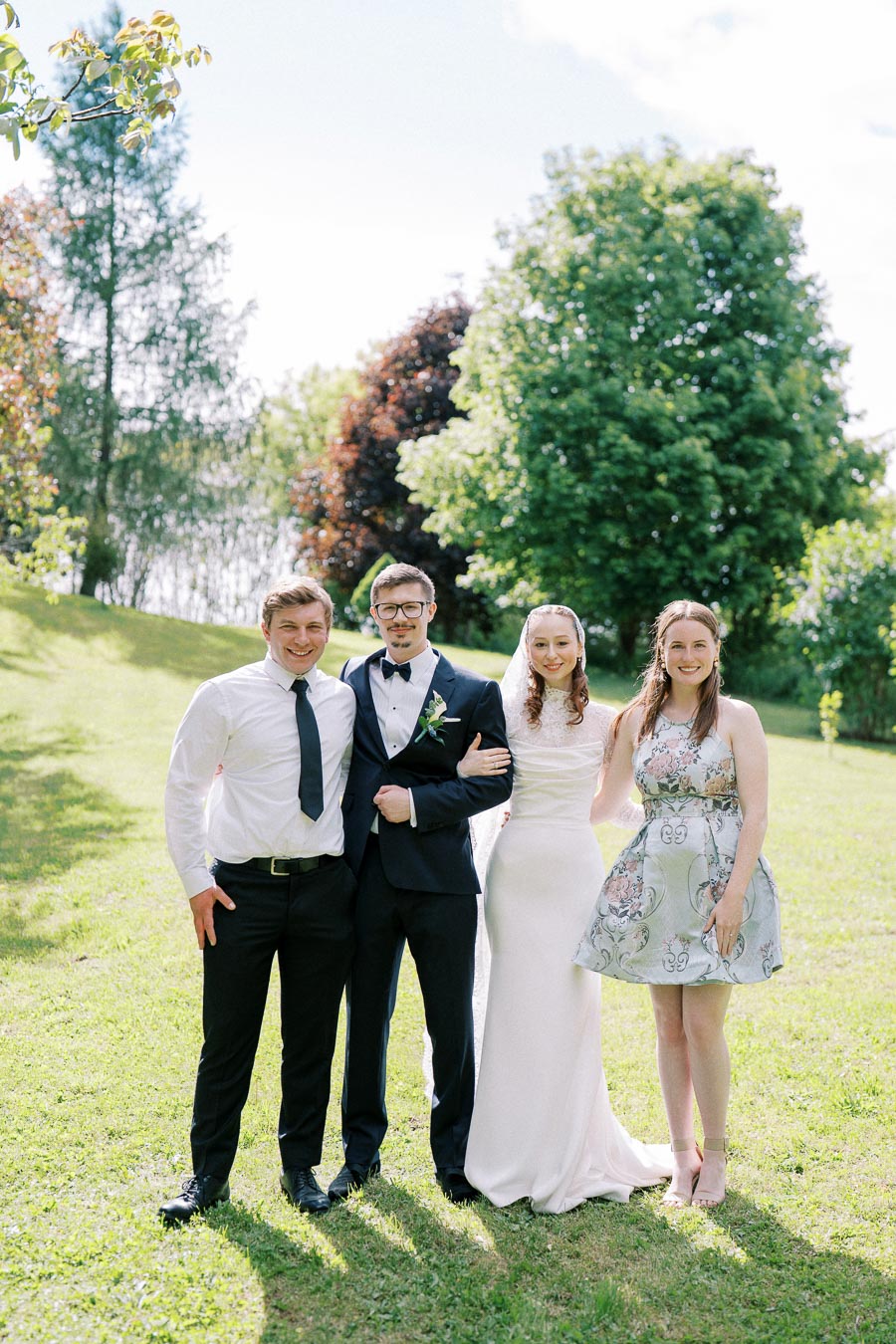 Outdoor wedding portrait with bride in white gown, groom in dark suit, and two guests posing on lush green lawn with trees in background.