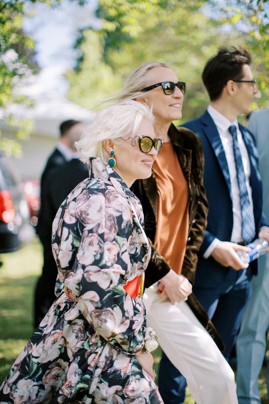 Elegant group of people attending an outdoor event, with individuals wearing fashionable outfits and sunglasses, standing in a garden with bright green surroundings.