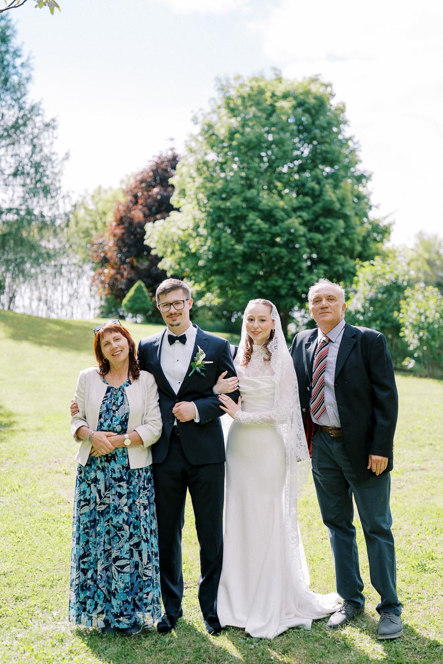 A joyful wedding scene featuring a bride in an elegant white gown and lace veil, standing beside the groom in a classic tuxedo. They are accompanied by two guests, all posing together on lush green grass with vibrant trees in the background, under a sunny sky.