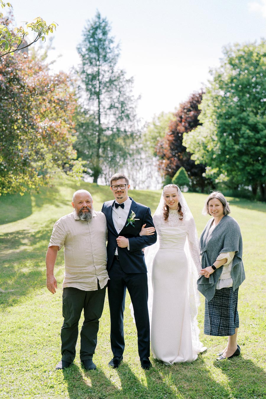 A joyful wedding scene in a sunlit garden with a groom and bride standing between an older couple, surrounded by lush greenery and a clear blue sky.