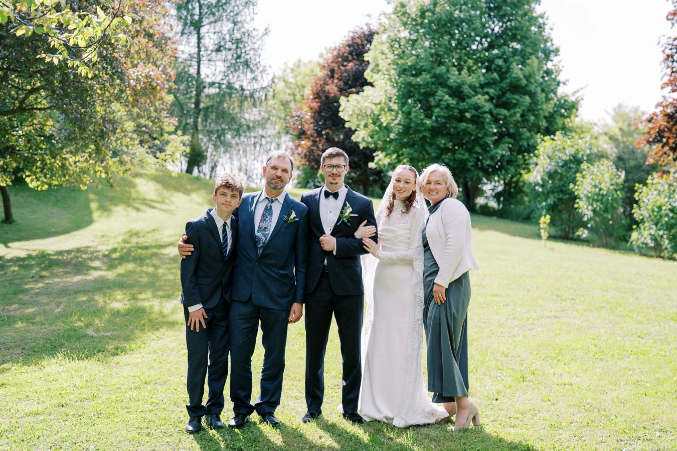 A group of people dressed in formal attire standing outdoors on a sunny day, surrounded by lush greenery and trees, capturing a joyful moment at a celebratory event.