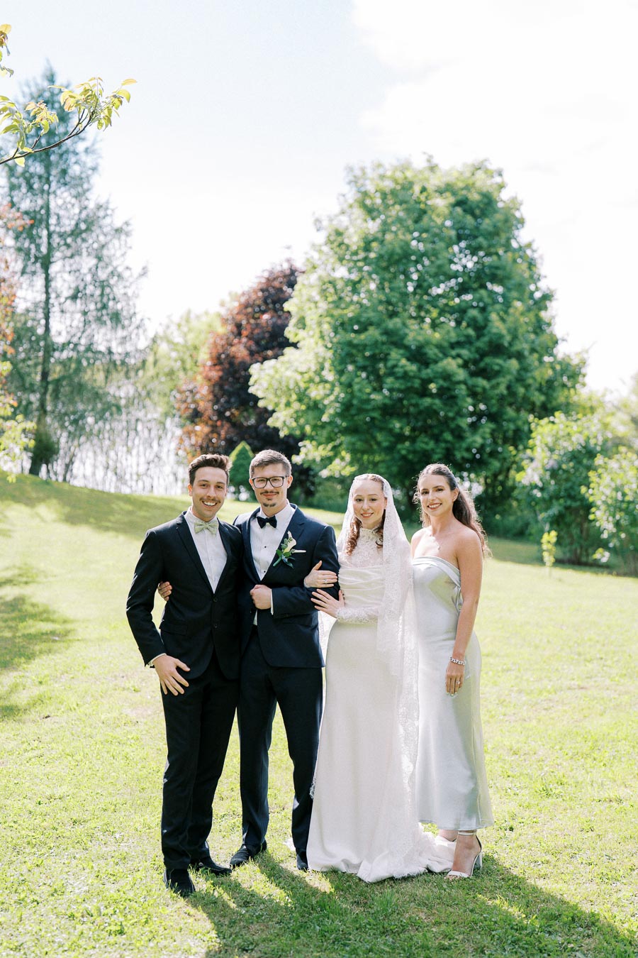 A group of four people posing for a wedding photo outdoors on a sunny day, with lush green trees in the background. The groom is in a tuxedo, the bride is wearing a white dress with a veil, while two family members or friends stand beside them smiling.