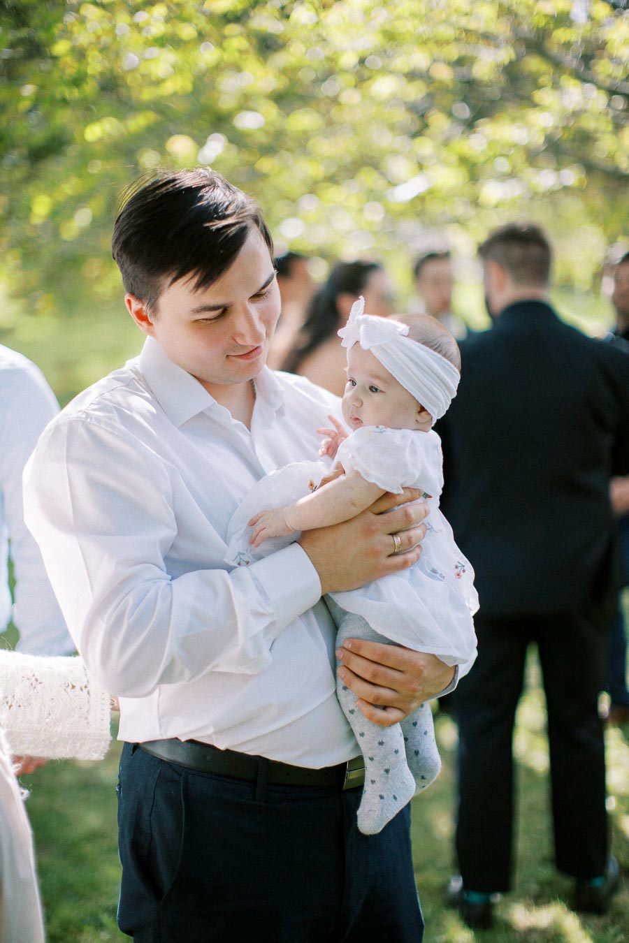 A man in a white shirt lovingly holds a baby dressed in white, wearing a headband, outdoors at a gathering with blurred people in the background.