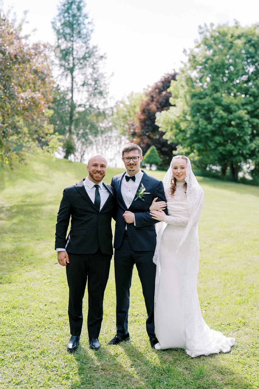 Three people posing outdoors on a sunny day; a woman in a white wedding dress, a man in a suit with a boutonniere, and another man in a suit, surrounded by lush greenery and trees.