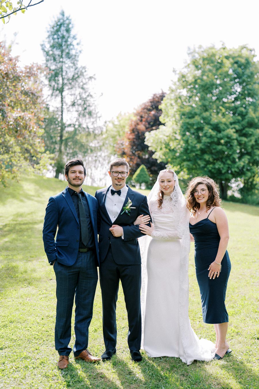A joyful wedding group photo captured outdoors on a sunny day, featuring a bride in a white gown, a groom in a black tuxedo, and two guests dressed in formal attire, with a lush green background of trees and grass.