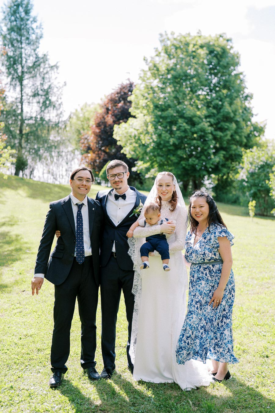 Outdoor wedding portrait featuring a smiling bride in a lace gown holding a baby, alongside her groom in a suit, and two guests, standing on a green lawn with trees in the background.
