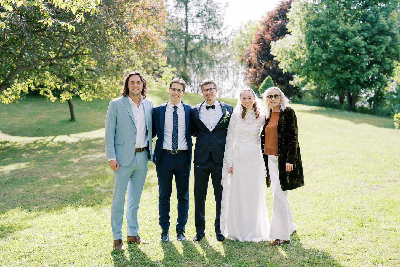 A group of five people standing together outdoors on a sunny day, dressed in formal attire. The scene features lush greenery and trees in the background, suggesting a celebration or event.