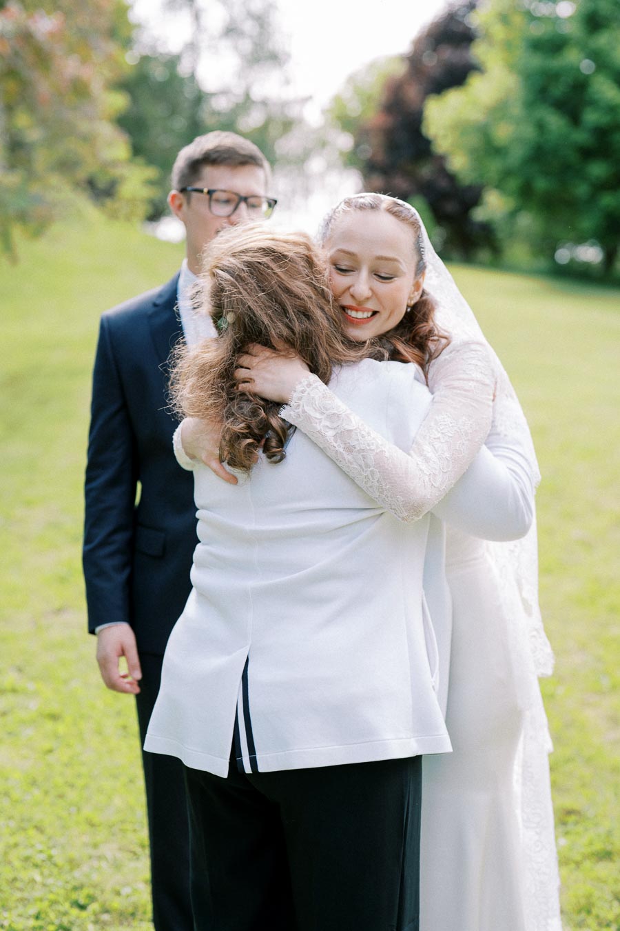A bride in a lace wedding dress embraces a guest during an outdoor wedding ceremony, with a man in a suit standing nearby on a grassy lawn.