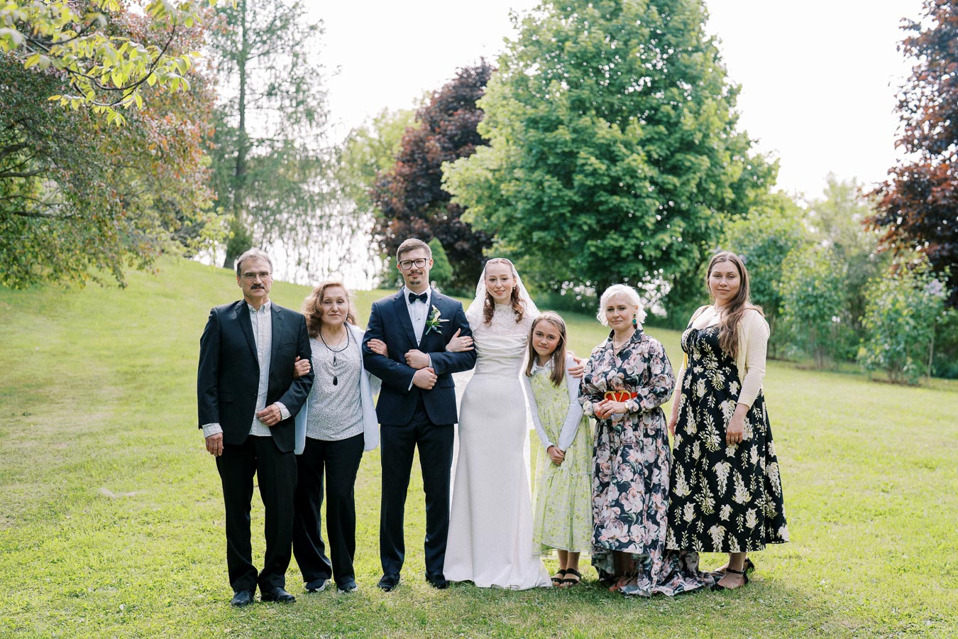 A wedding photo featuring a smiling bride in a white gown and groom in a dark suit surrounded by family members in a lush green park setting. The group poses in front of vibrant trees, capturing a joyful outdoor celebration.