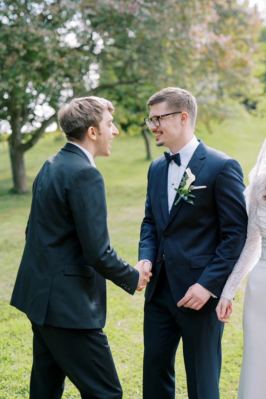 Two men dressed in formal suits cheerfully shaking hands outside, with a lush green background, capturing a joyful moment at an outdoor wedding.