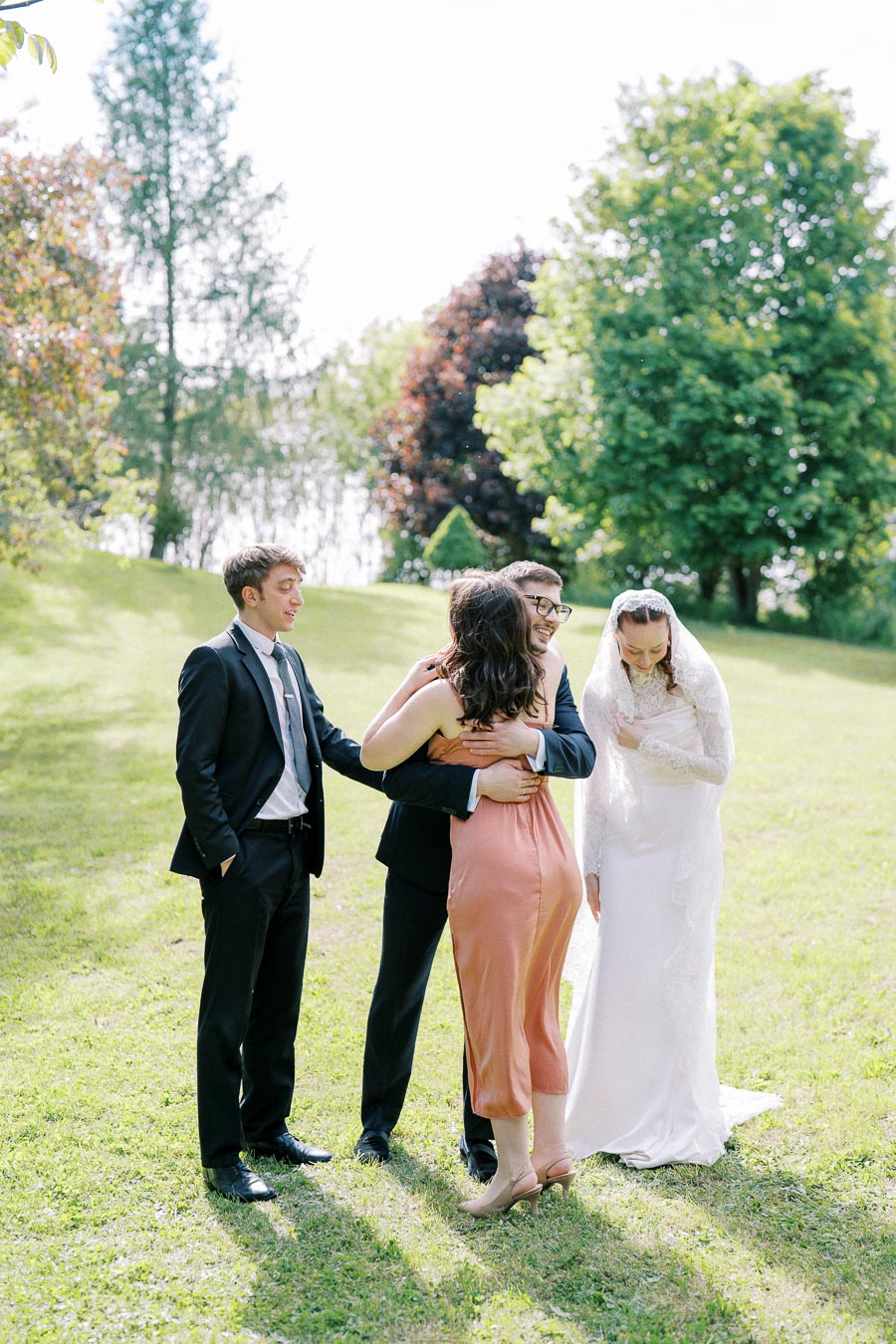 A joyful wedding scene capturing a bride in a white dress, a groom, and two guests in formal attire, sharing hugs and smiles on a lush green lawn under a bright sky.