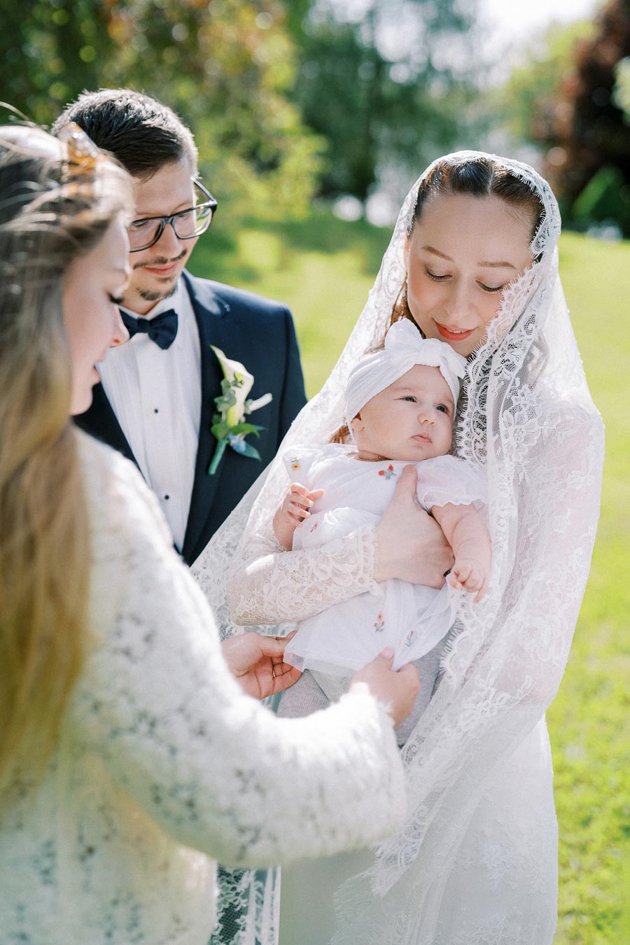 A family outdoor ceremony with a woman in a lace veil holding a baby dressed in white with a headband, a man in a suit and bow tie beside her, and another person reaching towards the baby. Lush greenery in the background.