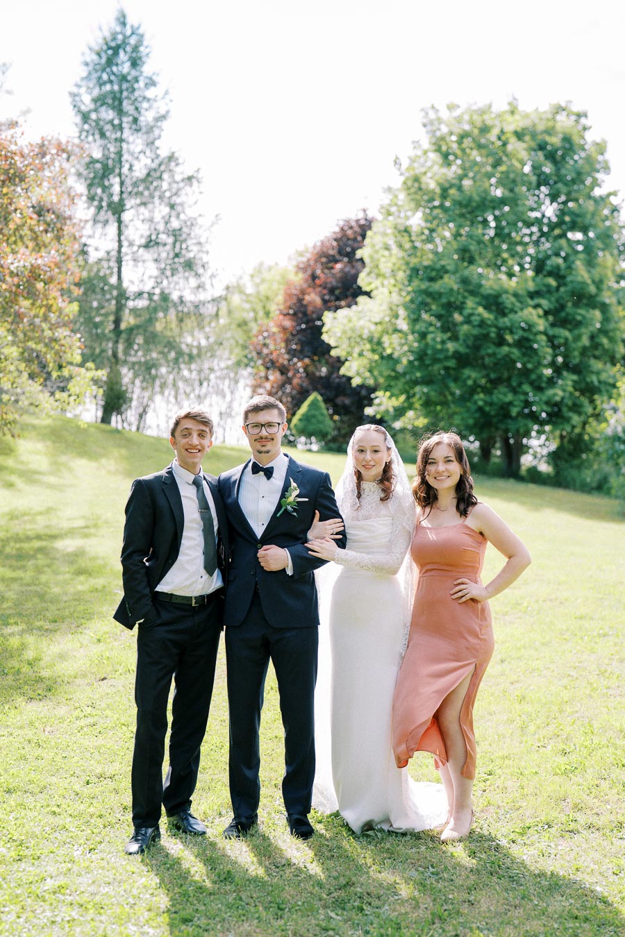 A wedding group photo featuring a bride in a white gown, a groom in a dark suit, and two guests in formal attire, posed on a lush green lawn with trees in the background on a sunny day.