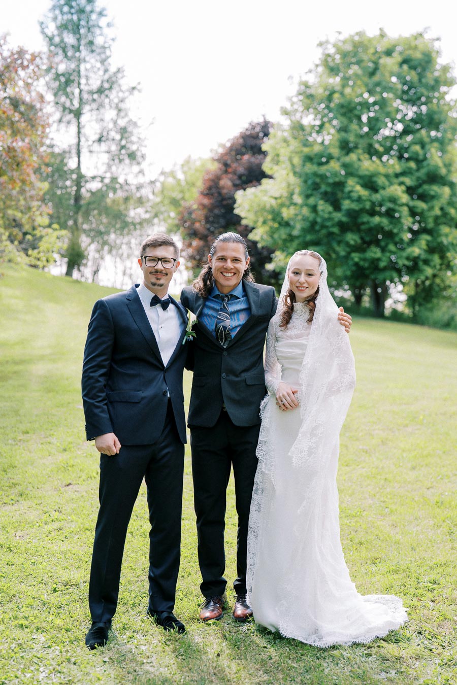 Three people standing together on a grassy field, with one wearing a wedding dress and veil, the others in suits, surrounded by lush green trees.